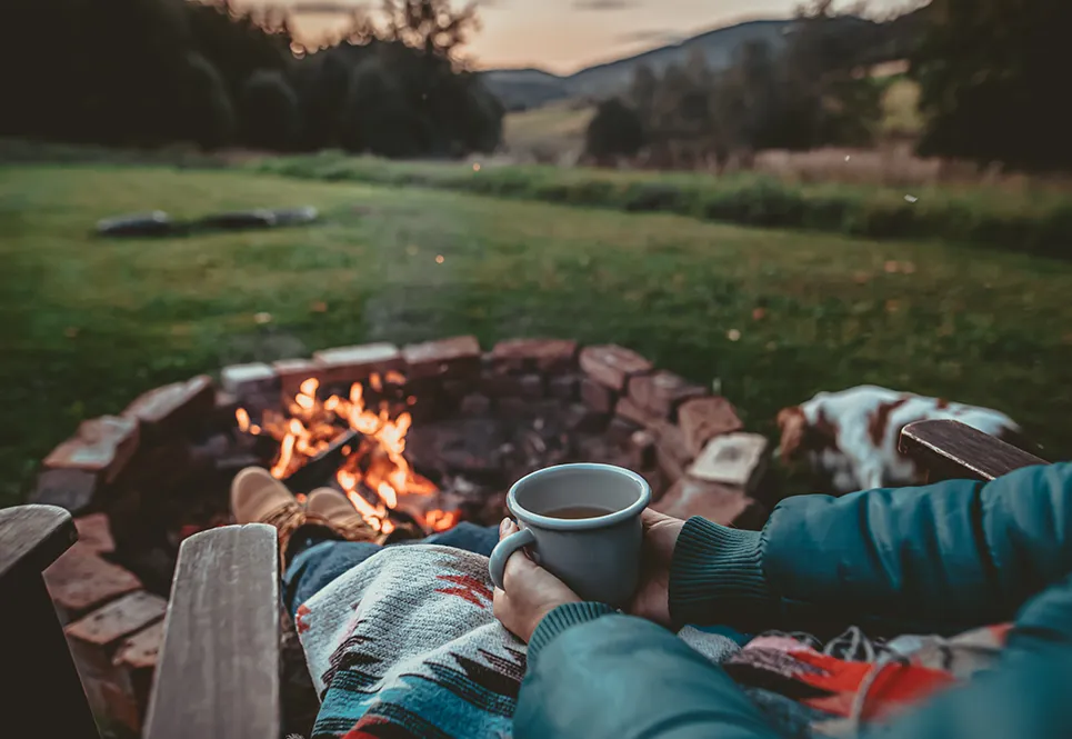 Person sitting outside, next to a warm fire, holding a mug of tea. 