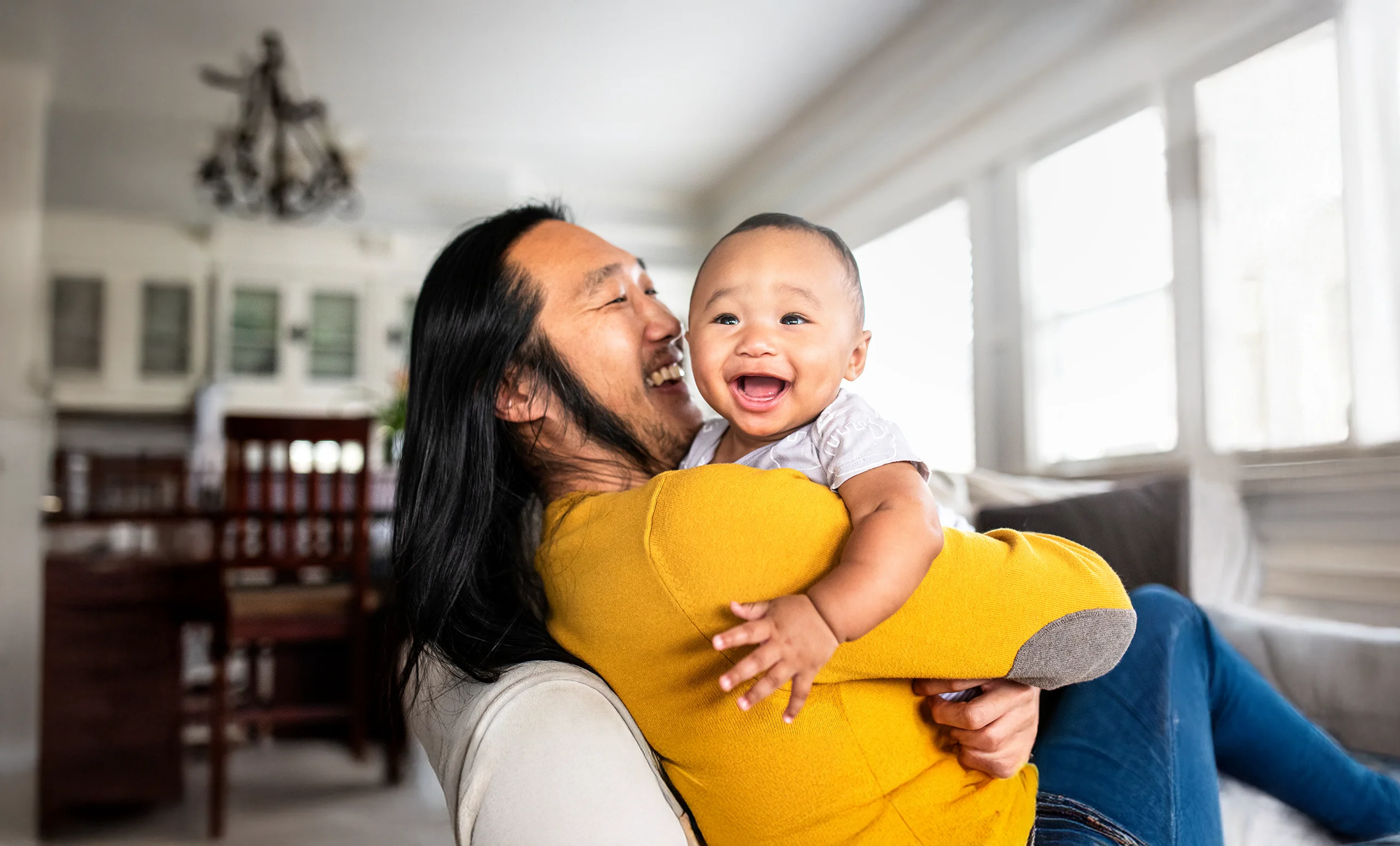 Smiling asian dad hugging his happy baby on a couch at home.