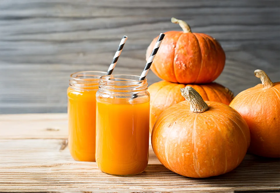 Two glasses of pumpkin juice with black and white striped straws next to pumpkins