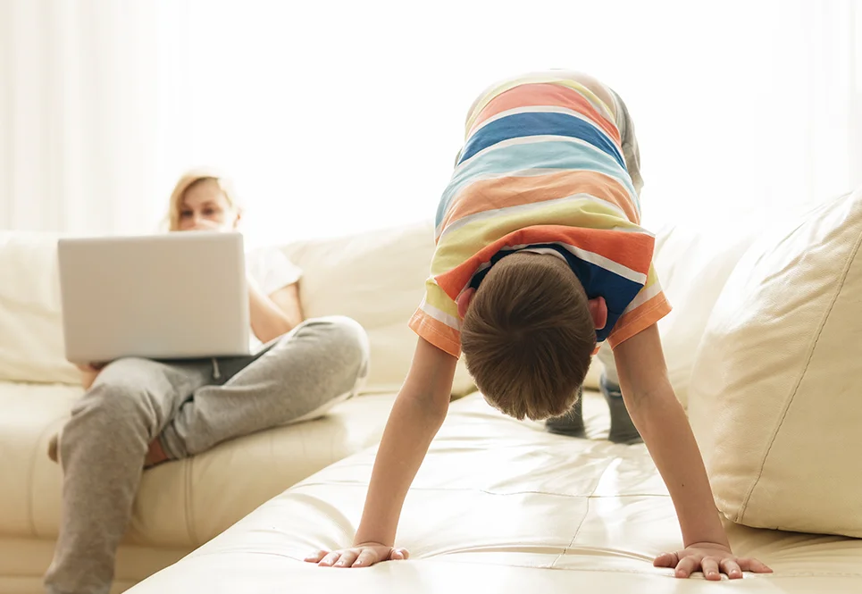 Young caucasian boy doing downward dog on a couch while mom is working