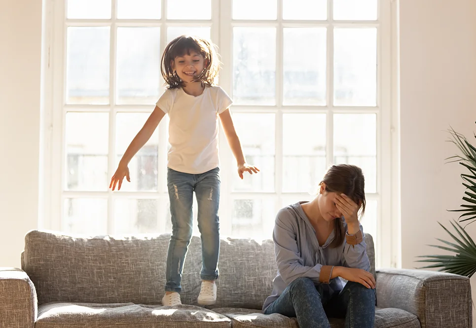 Little girl jumping on the sofa while her mom sits on it appearing upset