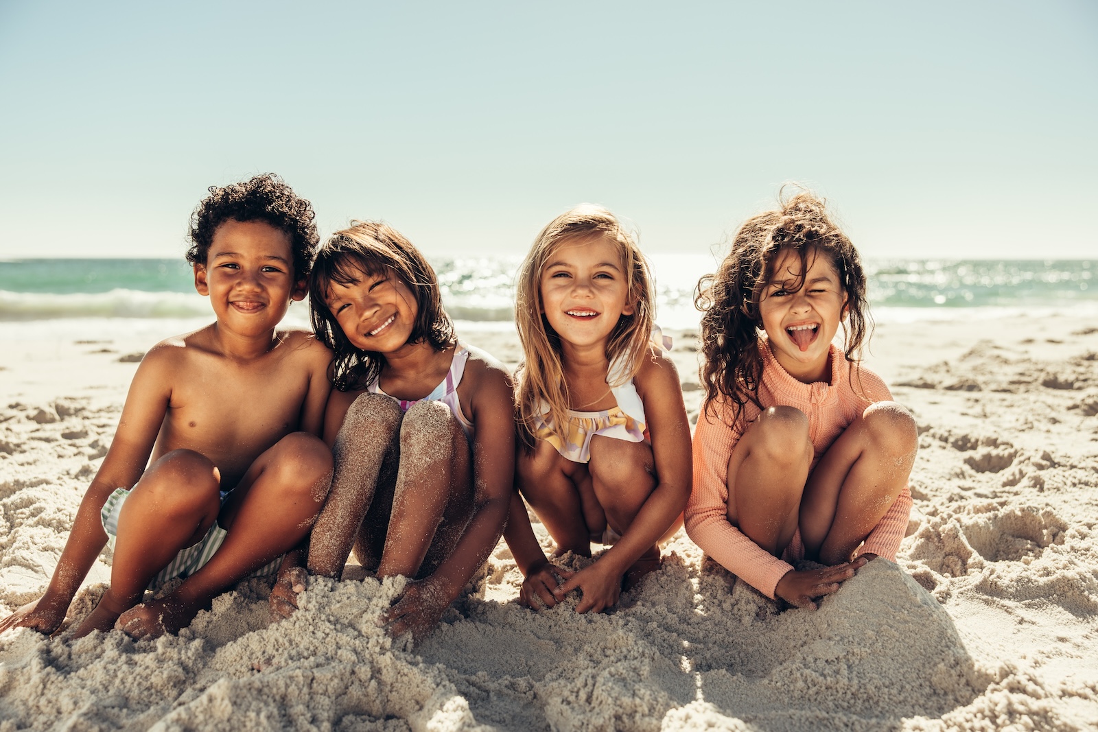 Cute little kids smiling at the camera while sitting in sea sand at the beach. Group of cheerful young children having a good time together during summer vacation.