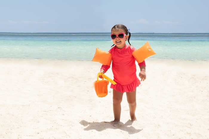 Portrait Of A Happy Toddler Girl Holding Plastic Toys In Hand Walking On Sandy Beach