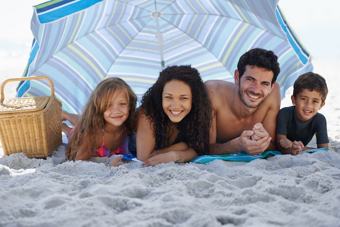 A happy family smiling at the camera while lying under an umbrella at the beach