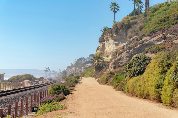 Train track and dirt road at the beach of San Clemente, California. There are rocky mountains with shrubs and trees on the right and a view of a sea on the left.