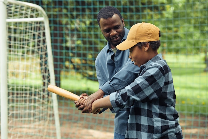 Black father helping his teen son swing a baseball bat. 