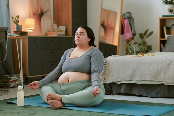 Pregnant woman doing yoga in her room, sitting cross legged on the floor. 