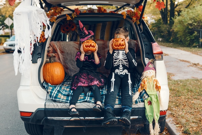 Two kids holding jack-o-lanterns in the open trunk of a car on Halloween