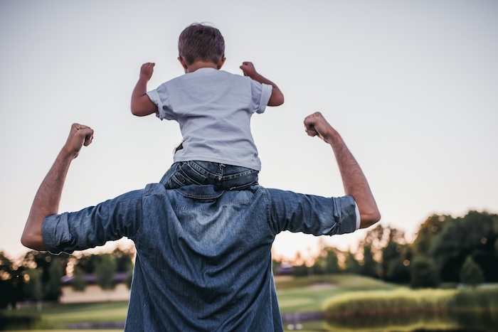 Child flexing his muscles, resting on his dad’s shoulders. 