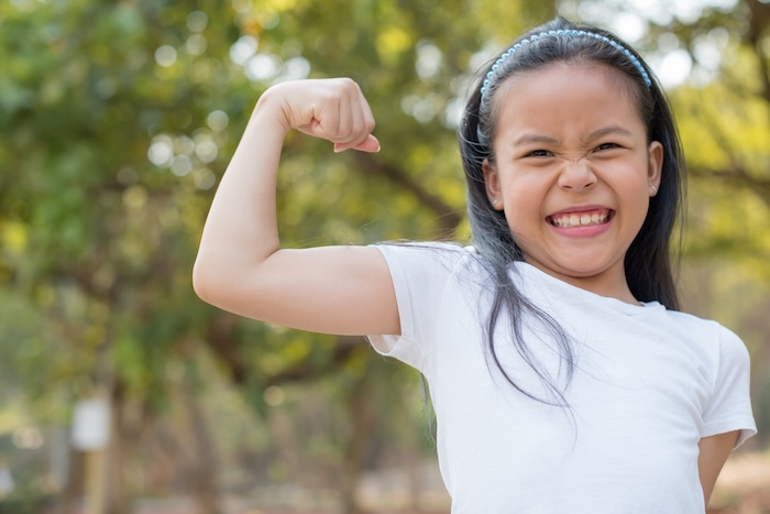 Young Asian child flexing her muscles outside in a green space. 