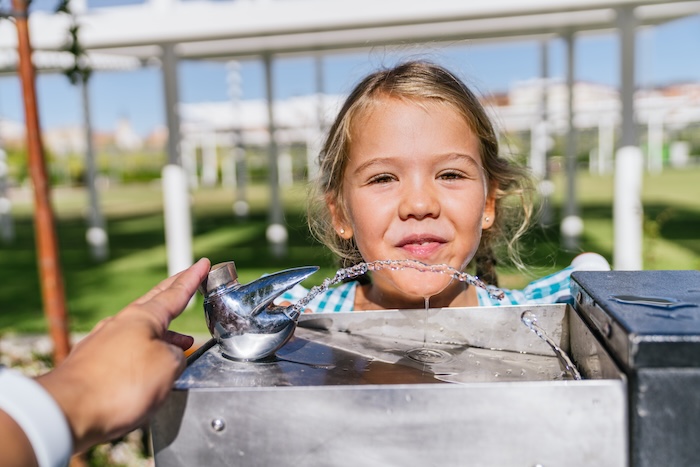 Young blonde girl drinking from a public water fountain on a hot summer day.