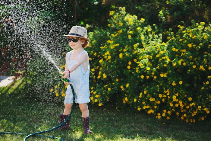 Young Caucasian boy playing with a water hose outside in a garden on a hot summer day. 
