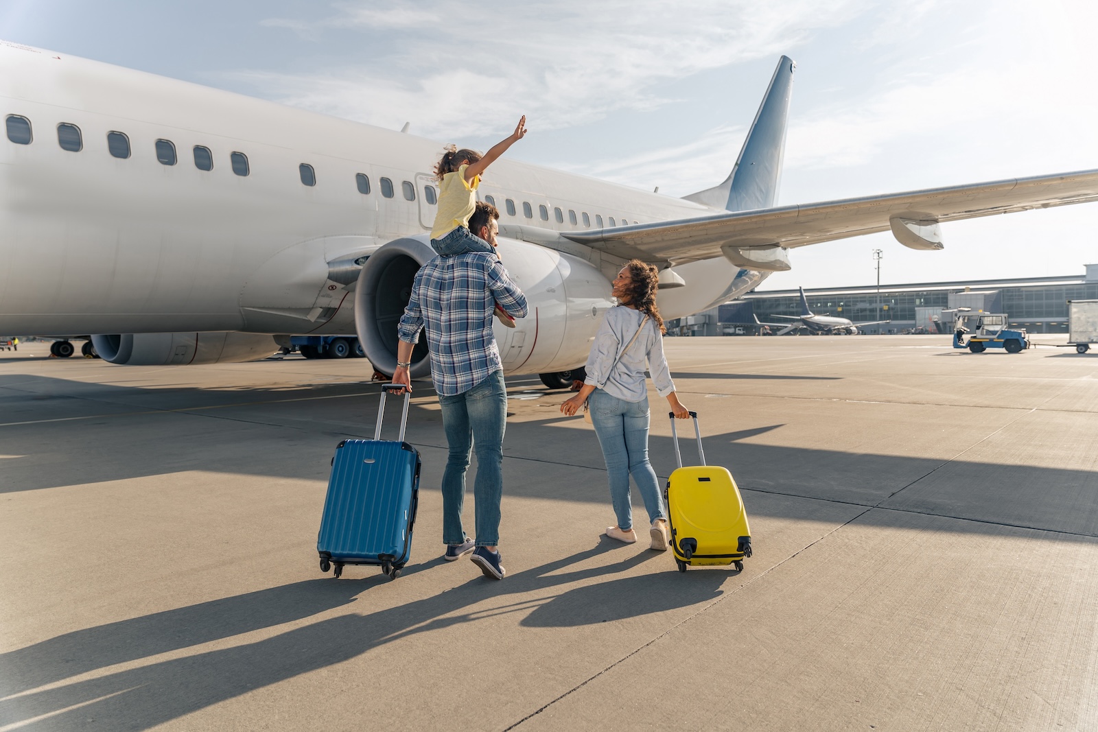 Caucasian family with a little girl on her dad’s shoulders on a tarmac to board a plane.