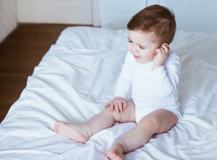 Toddler sitting on the bed, touching his ear in pain. 